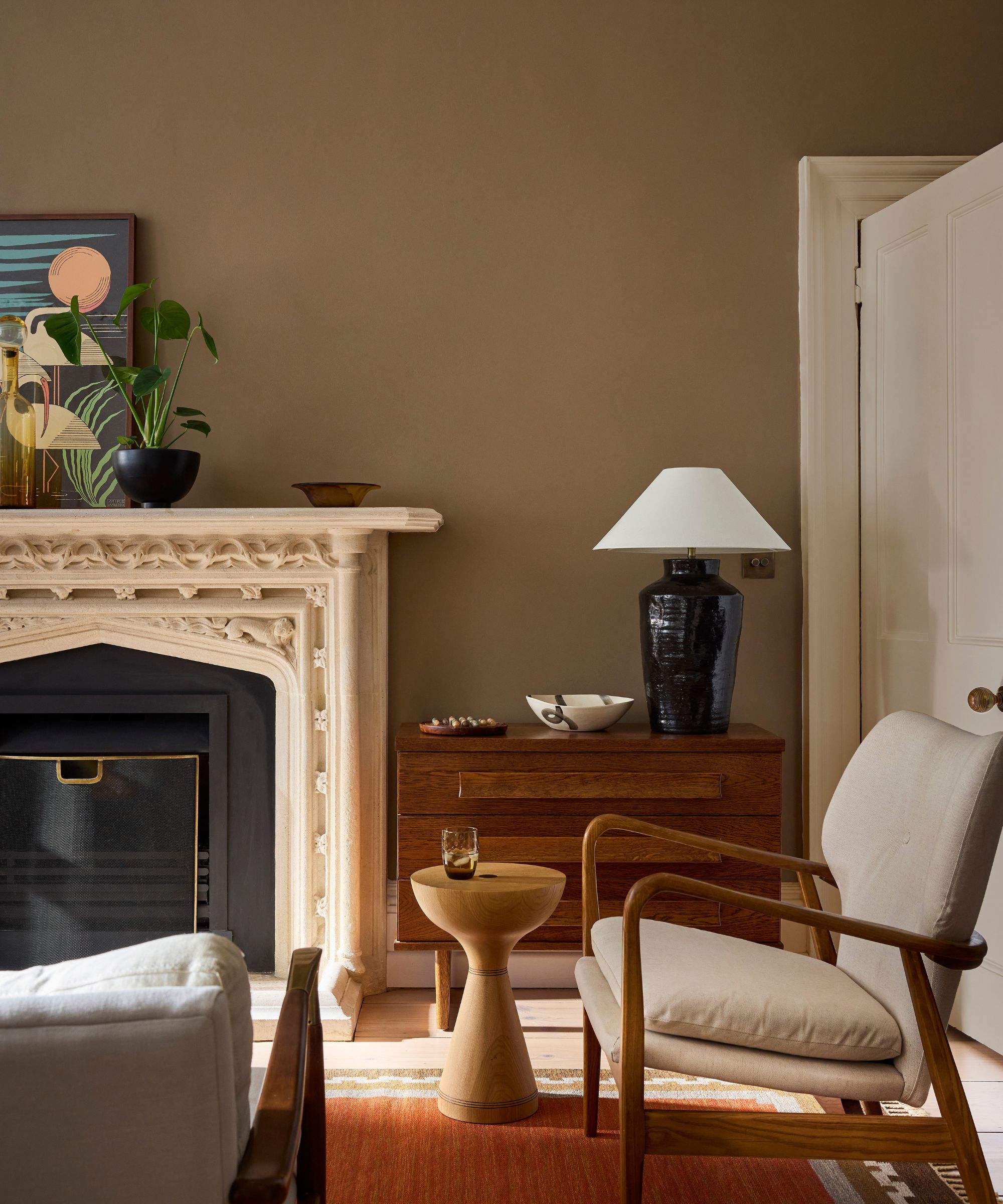 Living room with a marble fireplace, walls covered in Little Greene marble, a low-slung midcentury armchair, an orange rug, and a mahogany dresser topped with a large ceramic table lamp.