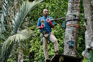 DI Mervin Wilson (Don Gilet) stands in the treetops, attached to a zipwire harness. He is on a platform, holding on to the wire and looking nervous.