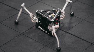 a black prism-shaped robot with three metal grey legs like a spider.