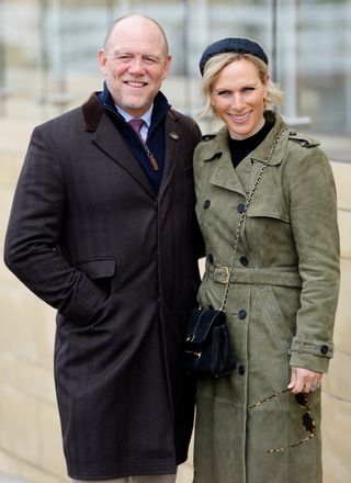 Mike Tindall wearing a dark coat with his hand in his pockets and Zara Tindall wearing a green coat and black headband smiling at the camera