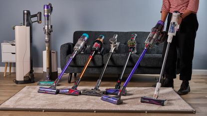 Writer holding two cordless stick vacuums, with 6 more resting on a sofa, and a carpeted panel on the floor