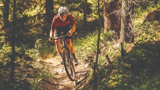A gravel rider rides a forested single track descent on a gravel bike