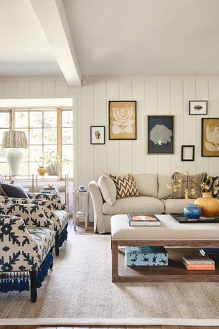 living room with yellow and blue accents, pair of blue matching armchairs, cream couch, ottoman with upholstered top, shiplap walls, artwork, vases, table lamps