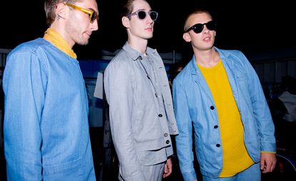 Male models wearing the Oliver Spencer S/S 2015 collection. All three models are wearing sunglasses The models on the left and right are wearing denim and mustard. The model in the middle is wearing a gray shirt, gray jacket and gray pants.