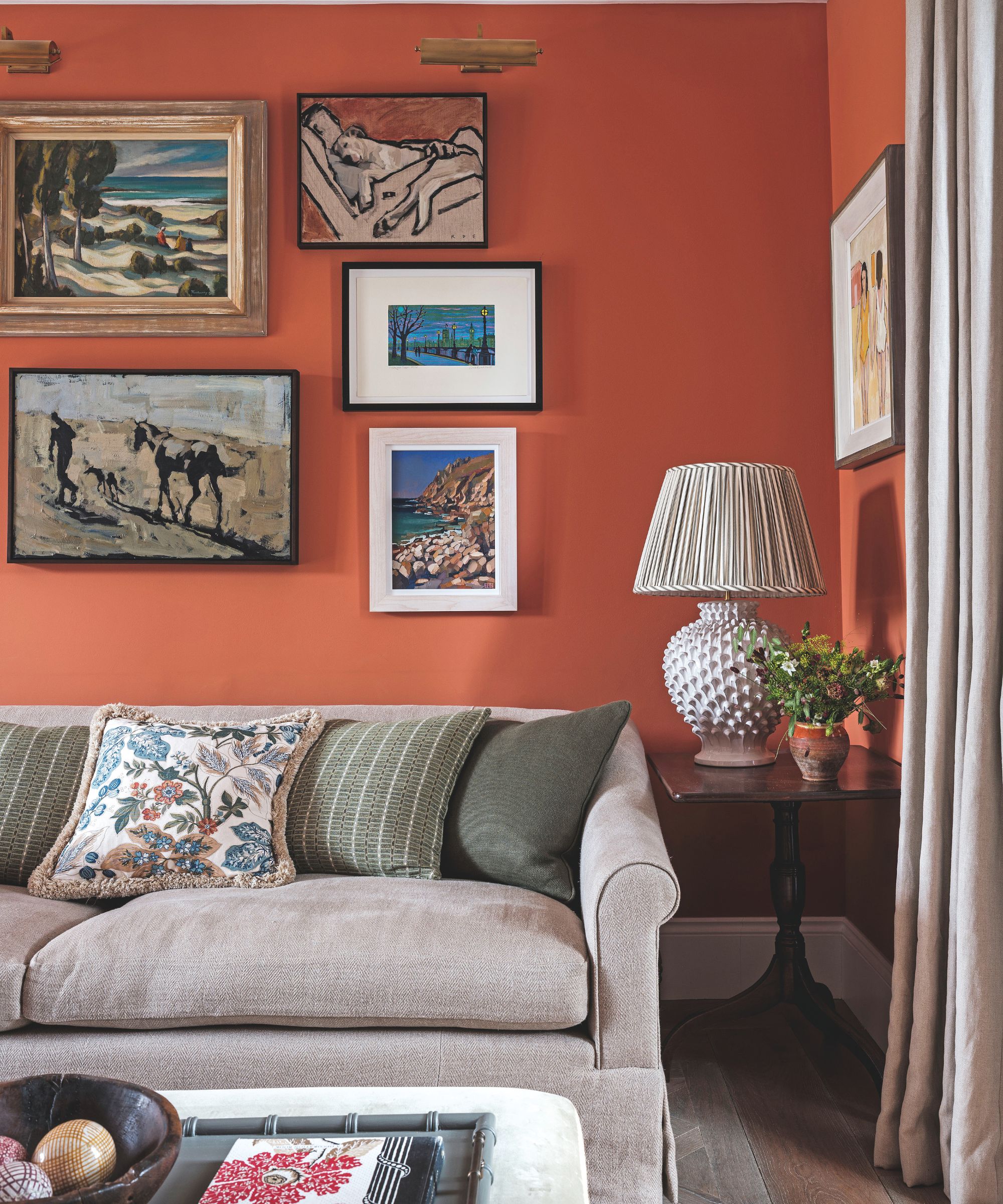 A cozy living room corner with a terracotta-colored wall decorated with a gallery of framed paintings above a neutral sofa with green cushions.