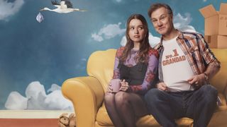 Aimee Lou Wood in a dark denim dress as Gemma sits on a yellow sofa next to David Morrissey in a No 1 Grandad t-shirt as a cartoon stork flies behind them in Daddy Issues