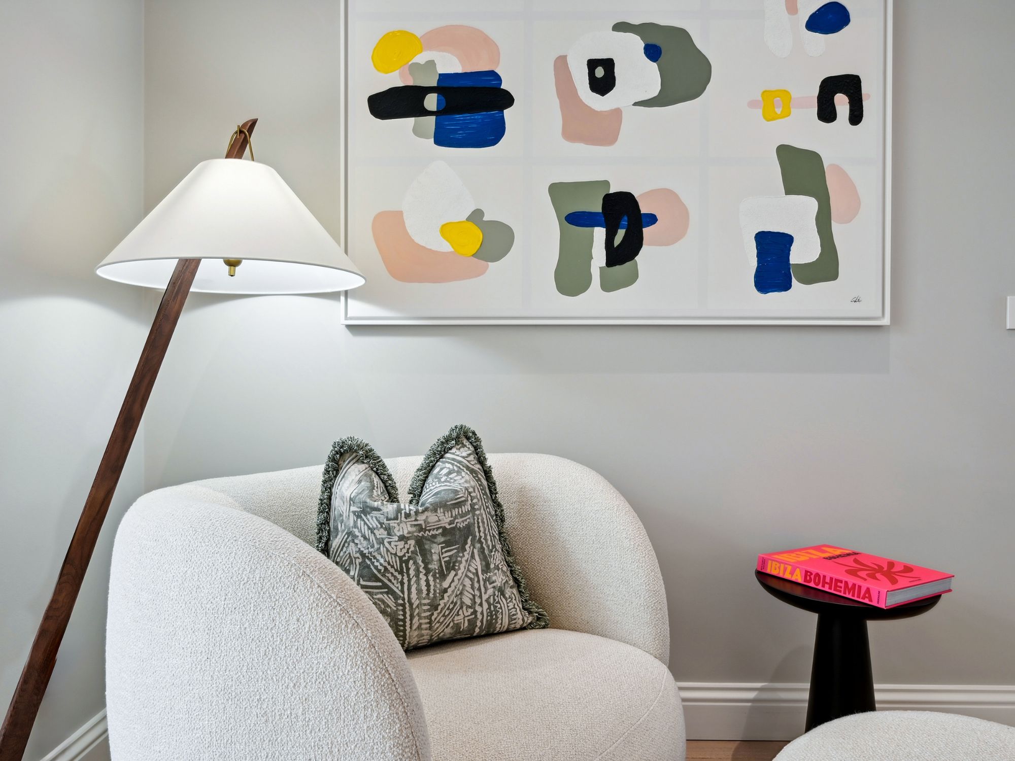 White and grey cozy minimalist home with lighting, boucle armchair, printed cushion, black table, pink book and abstract art on the wall