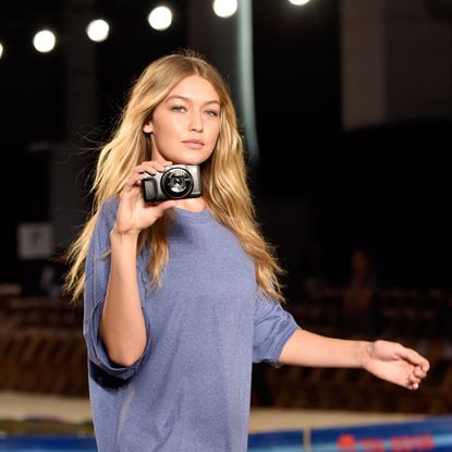 Gigi Hadid walking while pointing a compact camera at the person taking her picture.