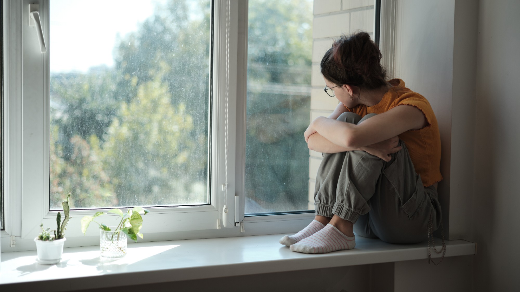 woman in yellow shirt and grey-green pants sits in a windowsill. she looks outside and might be sad or lonely