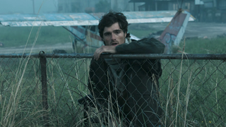 Jacob Elordi's Hig standing with arm on chain link fence while holding gun in other hand, with a plane in the backgroun, in The Dog Stars
