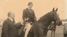 A sepia-toned image of a show pony ridden by Nigel Hollings, who has removed his hat to accept his prize, with the judge standing alongside, who has also removed his hat.