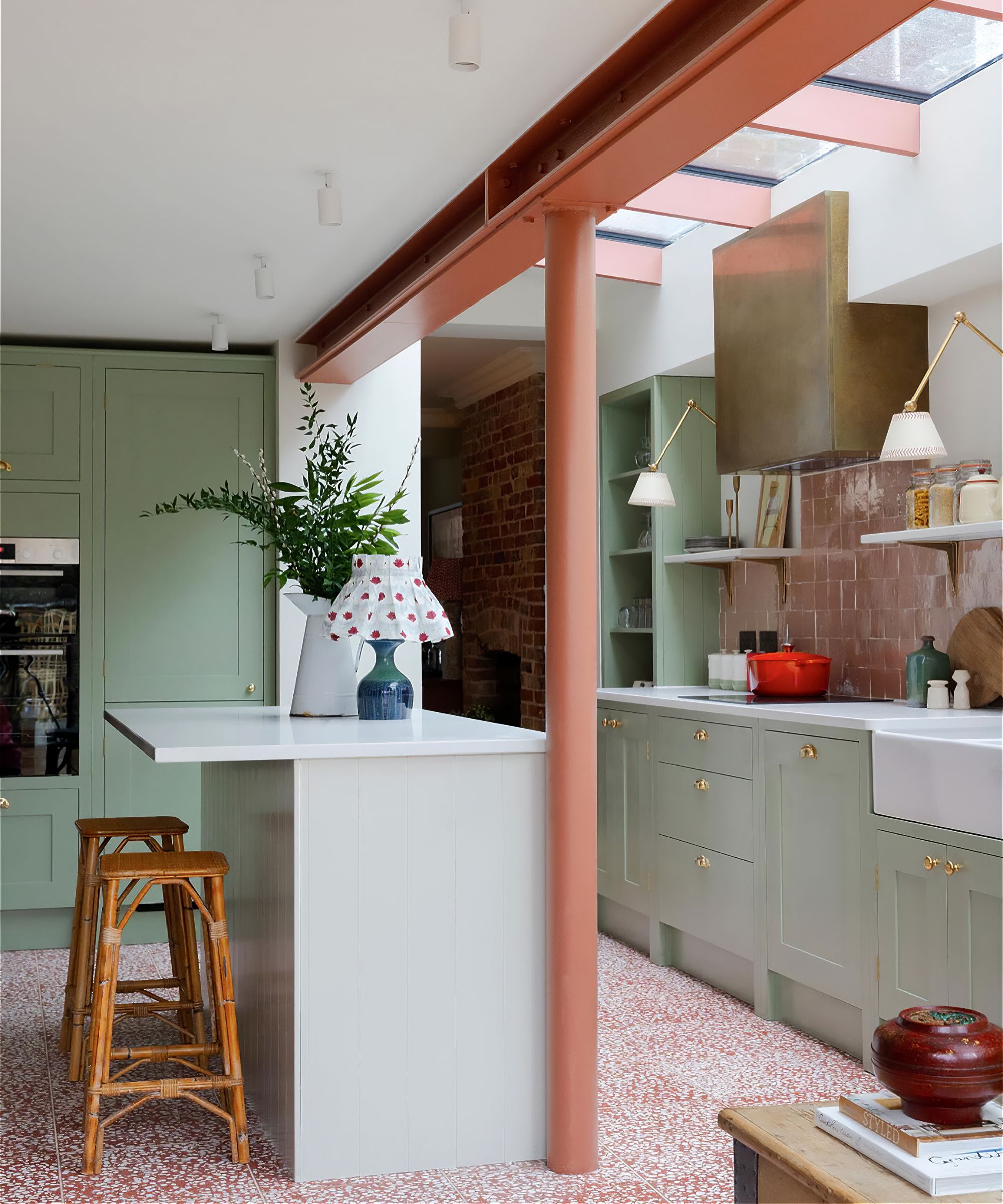 A sage and orange kitchen with exposed beams and a white kitchen island with wooden stools. To the right are sage green cabinets with a white sink, gold extractor fan, and a pink Zellige tiled backplash. The floor is red and white patterned tiling.