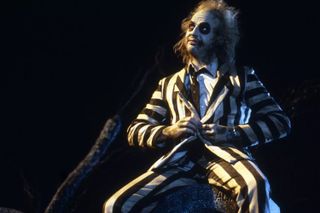 a man (michael keaton as beetlejuice) wearing a white-and-black striped suit and face makeup sits on a headstone