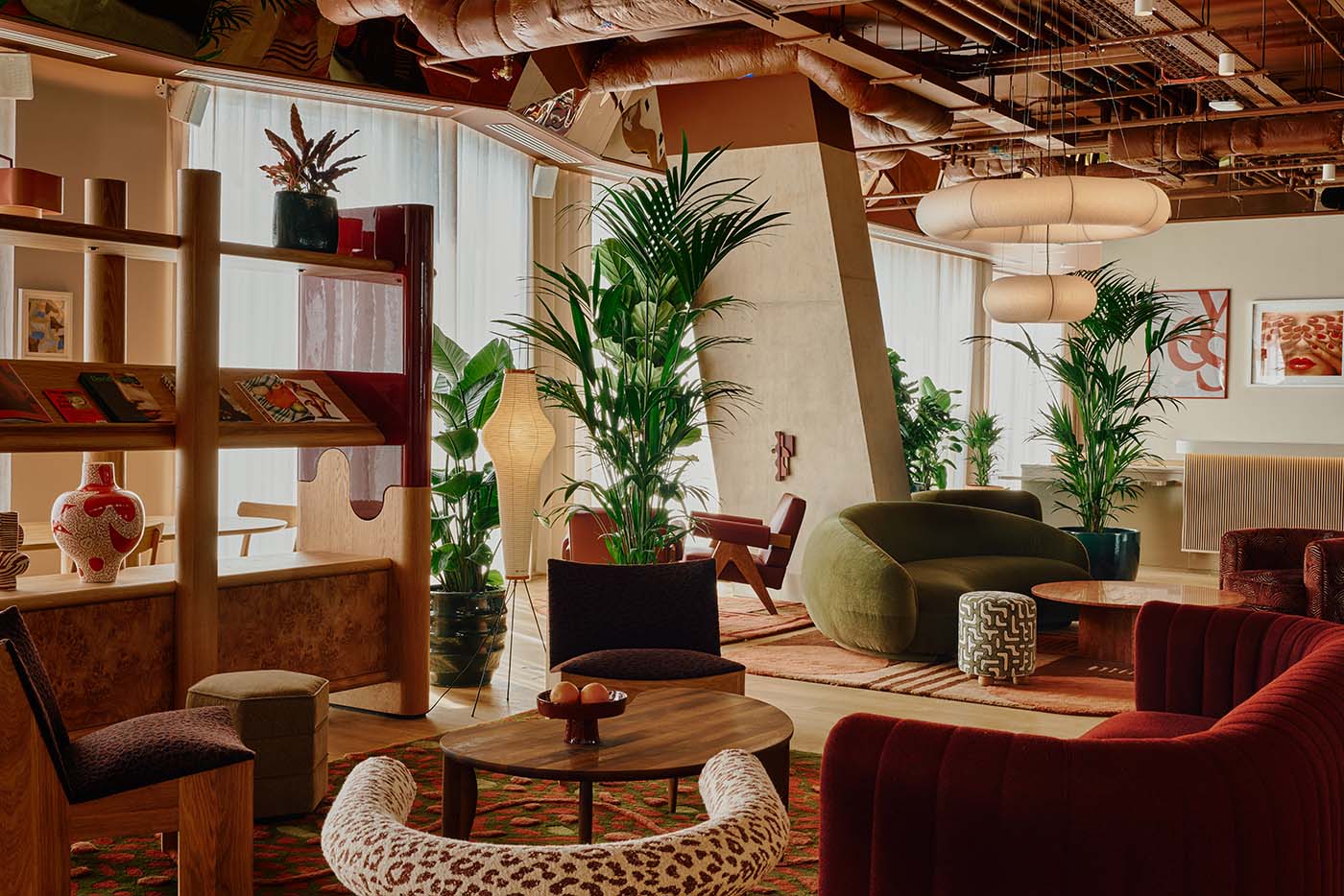 Interiors of a hotel lounge with red, green, and brown velvet sofas and armchairs, wooden furniture, thriving plants, industrial piping and uncovered ceilings, cement pillars, and warm lighting.