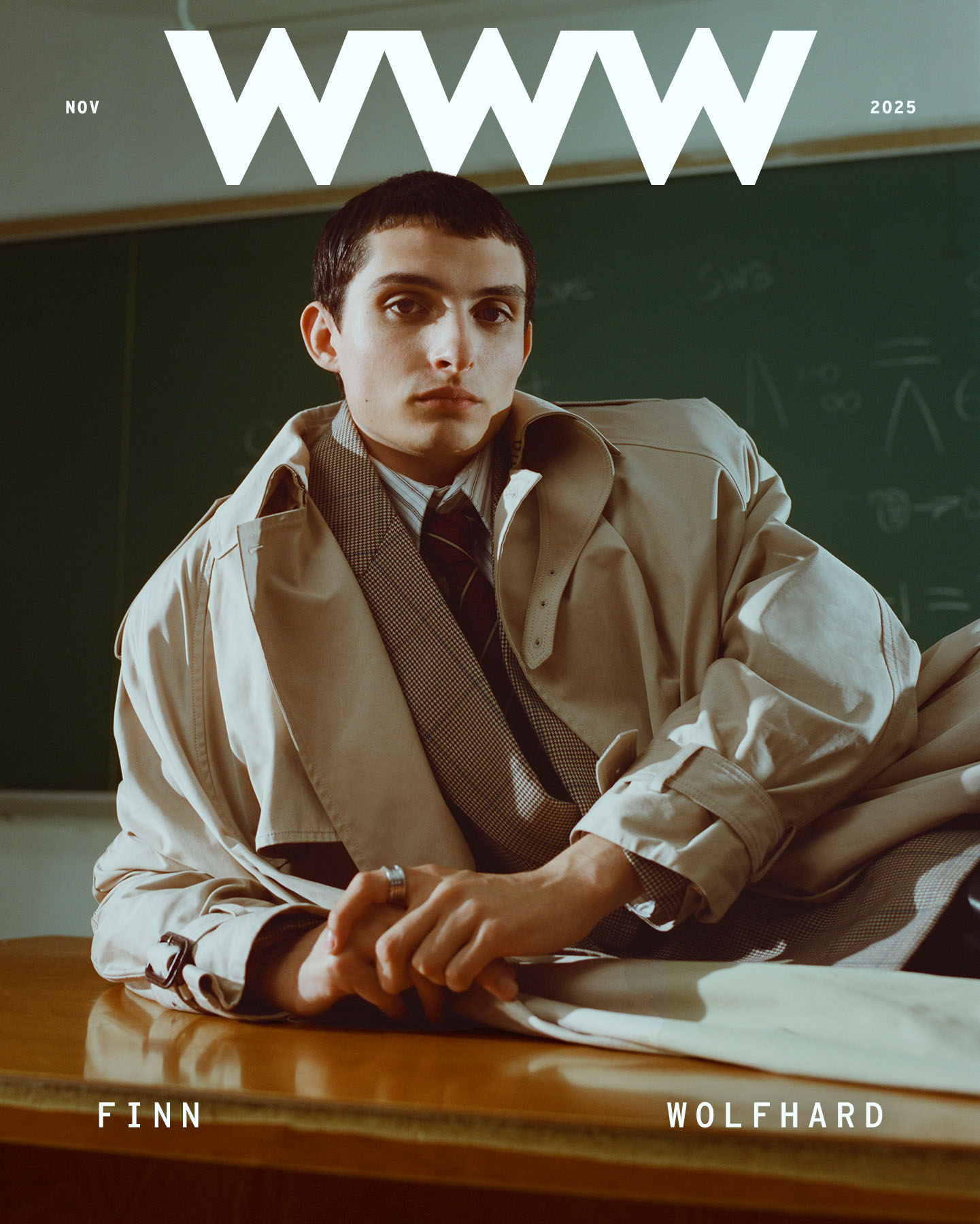 Finn Wolfhard wearing a beige trench coat, plaid blazer, striped shirt, and tie lying on a wooden desk.