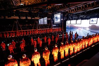A runway stage with men in orange construction workwear and hard hats lined up and facing different directions.