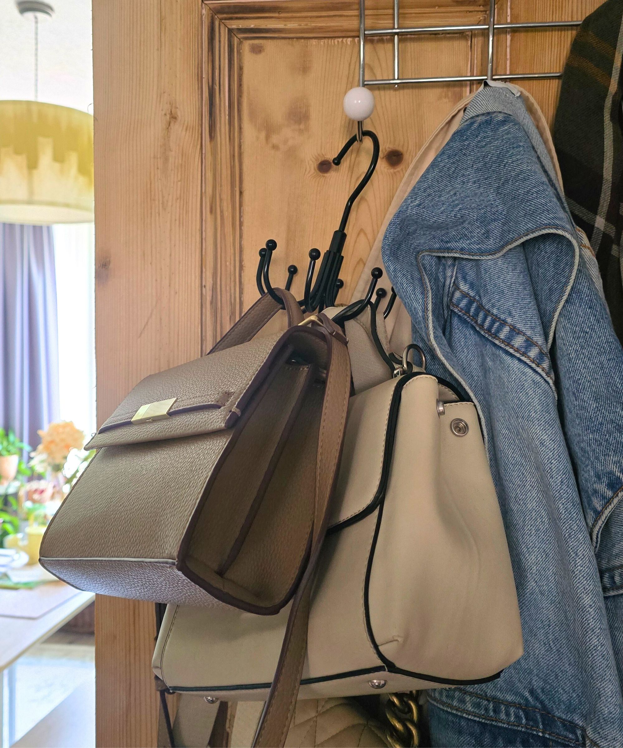 A wooden door with bags hanging from a black hook, coats hanging from a silver hook, and a green living room in the background