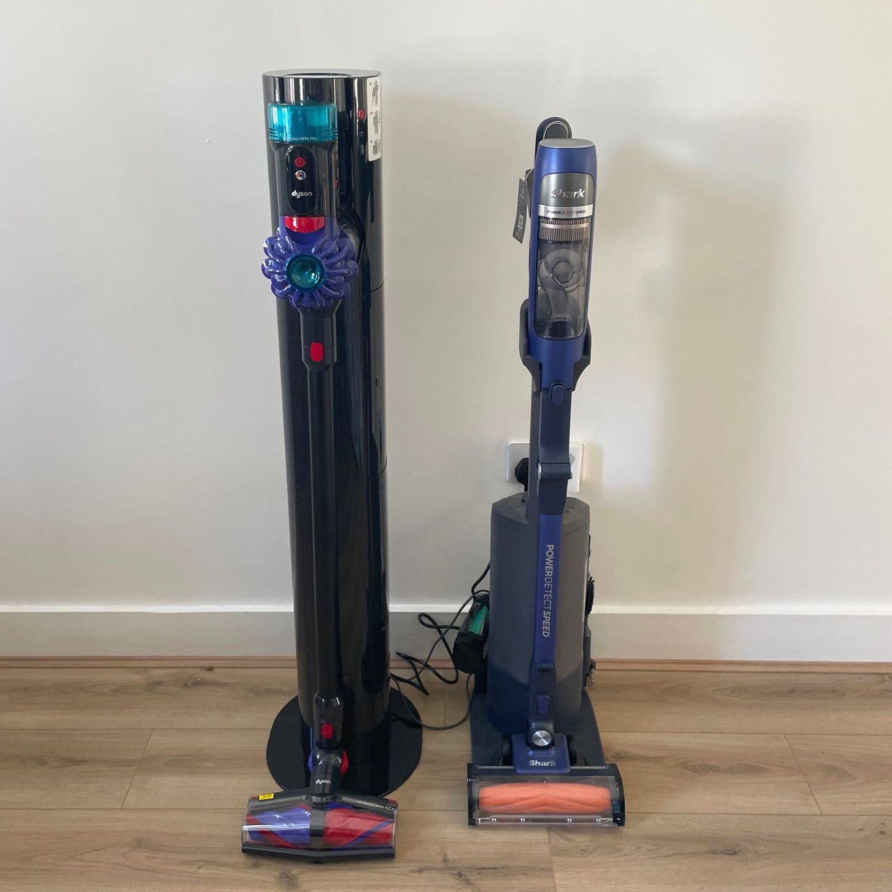 Dyson V10 Konical and auto-empty Dok standing next to the Shark PowerDetect Speed on wood-effect flooring, in front of a white wall