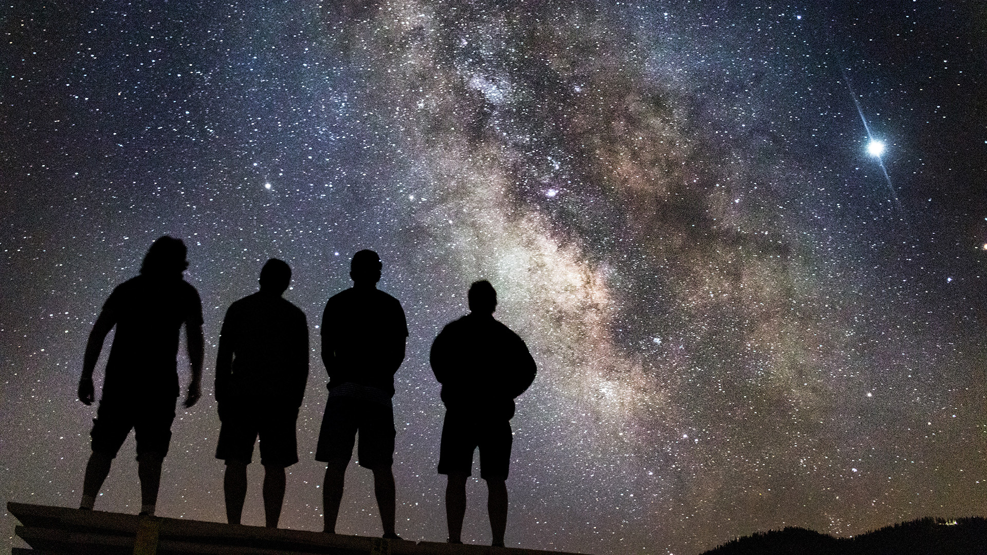 Men silhouetted in front of the night sky with the band of the Milky Way stretching across the sky.
