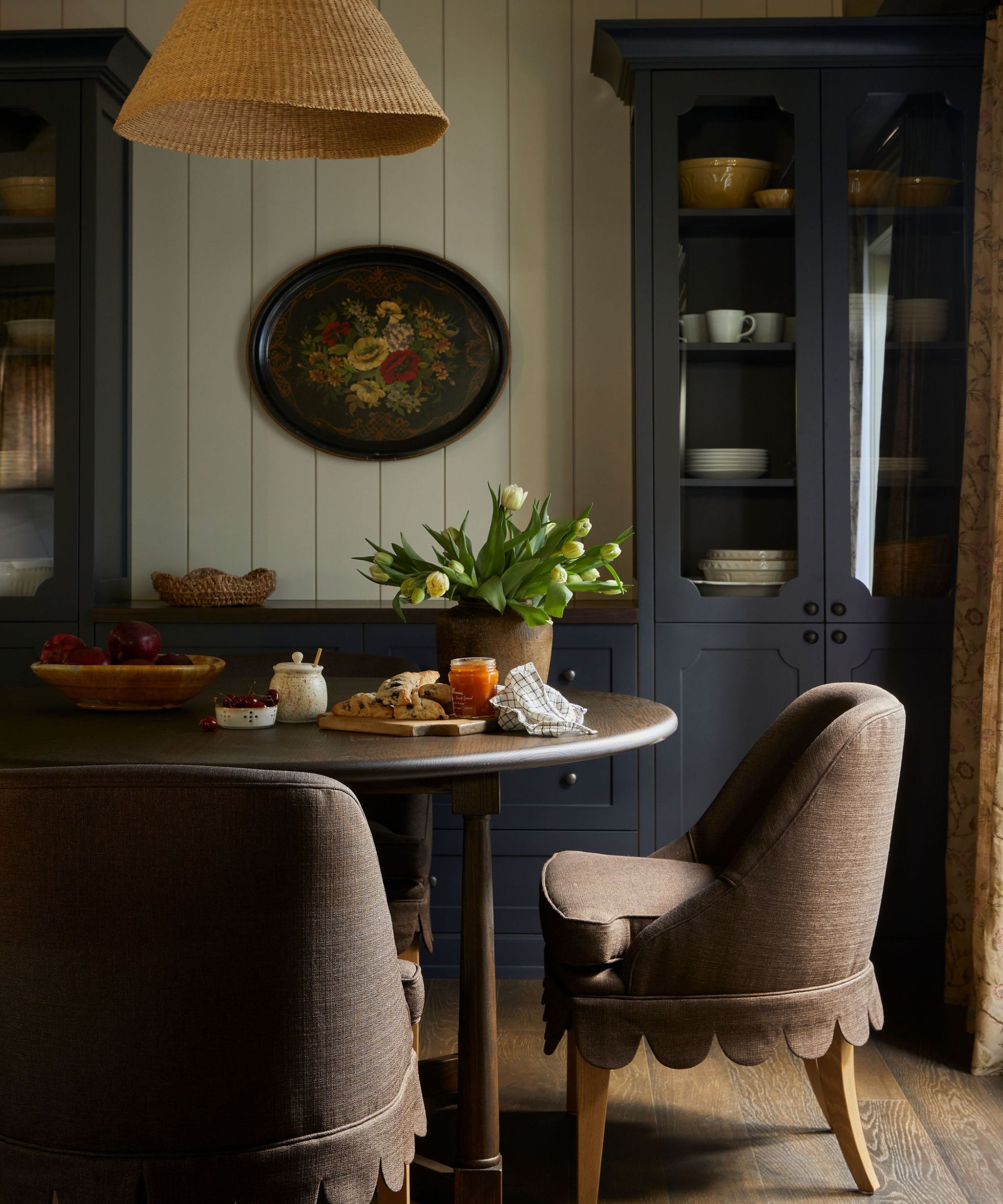Rustic dining room with built in blue cabinetry with glass-fronted doors, shelves decorated inside. Upholstered dining table around wooden table, decorated with flowers and a board of food