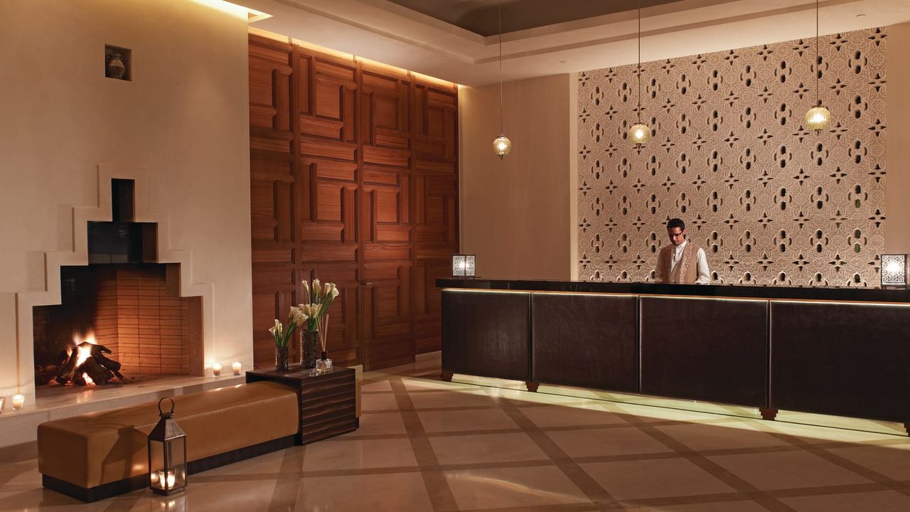 Image of the lobby of Four Seasons Marrakech. It is a plaster material with a large wooden wall and a fireplace.