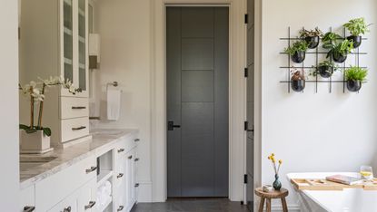 White bathroom with built-in cupboards, grey internal doors and plant grid on wall above white bath