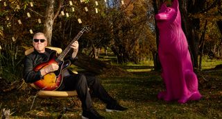 Larry DiMarzio wears shades and plays an ES-335 in a garden, under a tree decorated with his pickups. A giant pink plastic dog looks on.