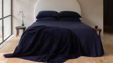 Dark navy merino jersey fitted bedding from SHLEEP on a large bed by a small wood bedside stool with a vase