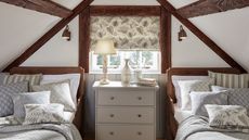 A cozy attic bedroom with two beds on either side of a small window. A white painted dresser between them