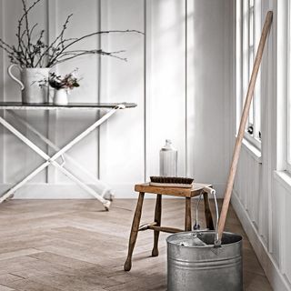 Zinc bucket and mop on wooden floor, in a room with a foldable white table with brown top against a white wall