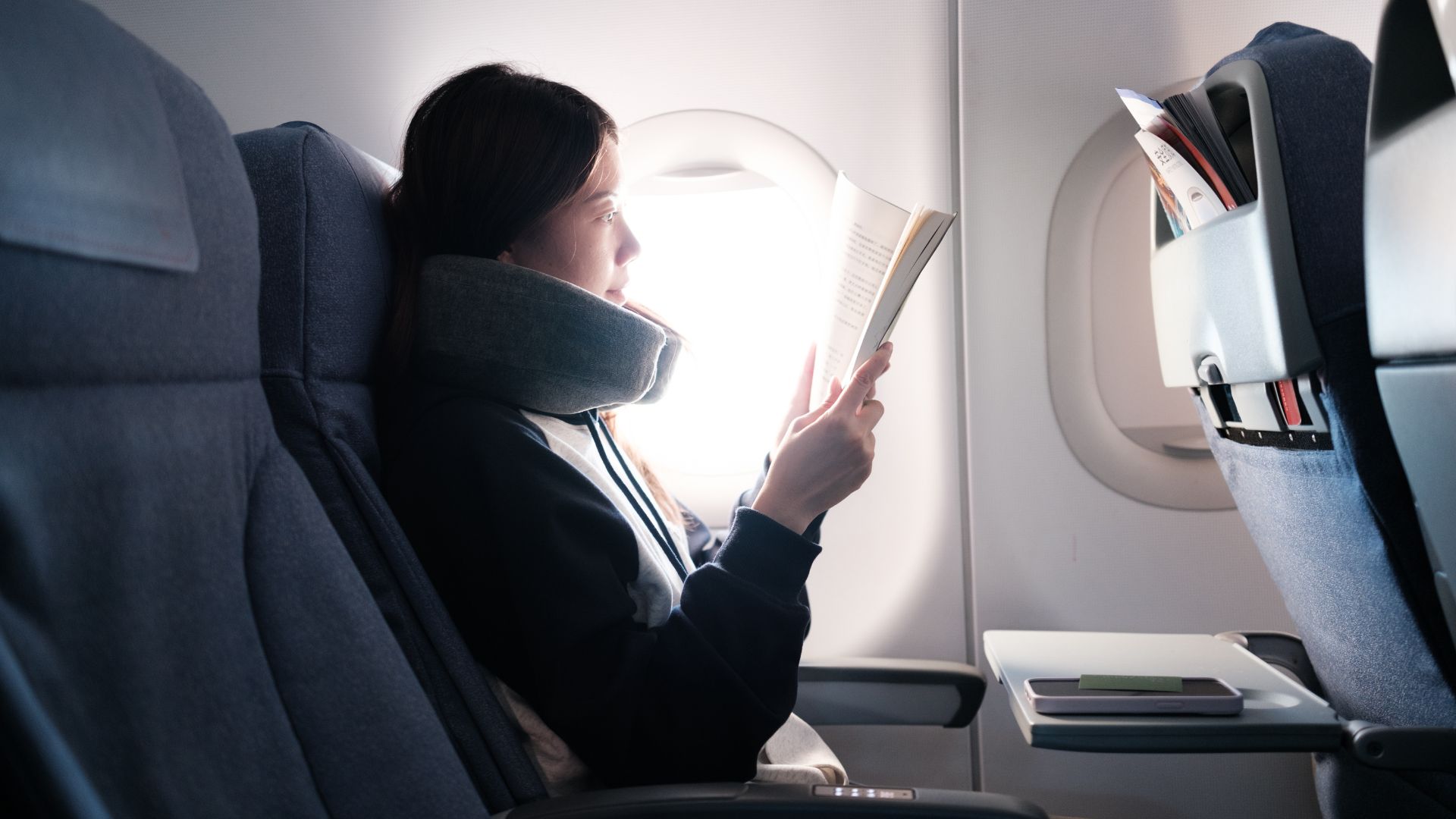 A woman reading a book in window seat of a plane with a neck pillow