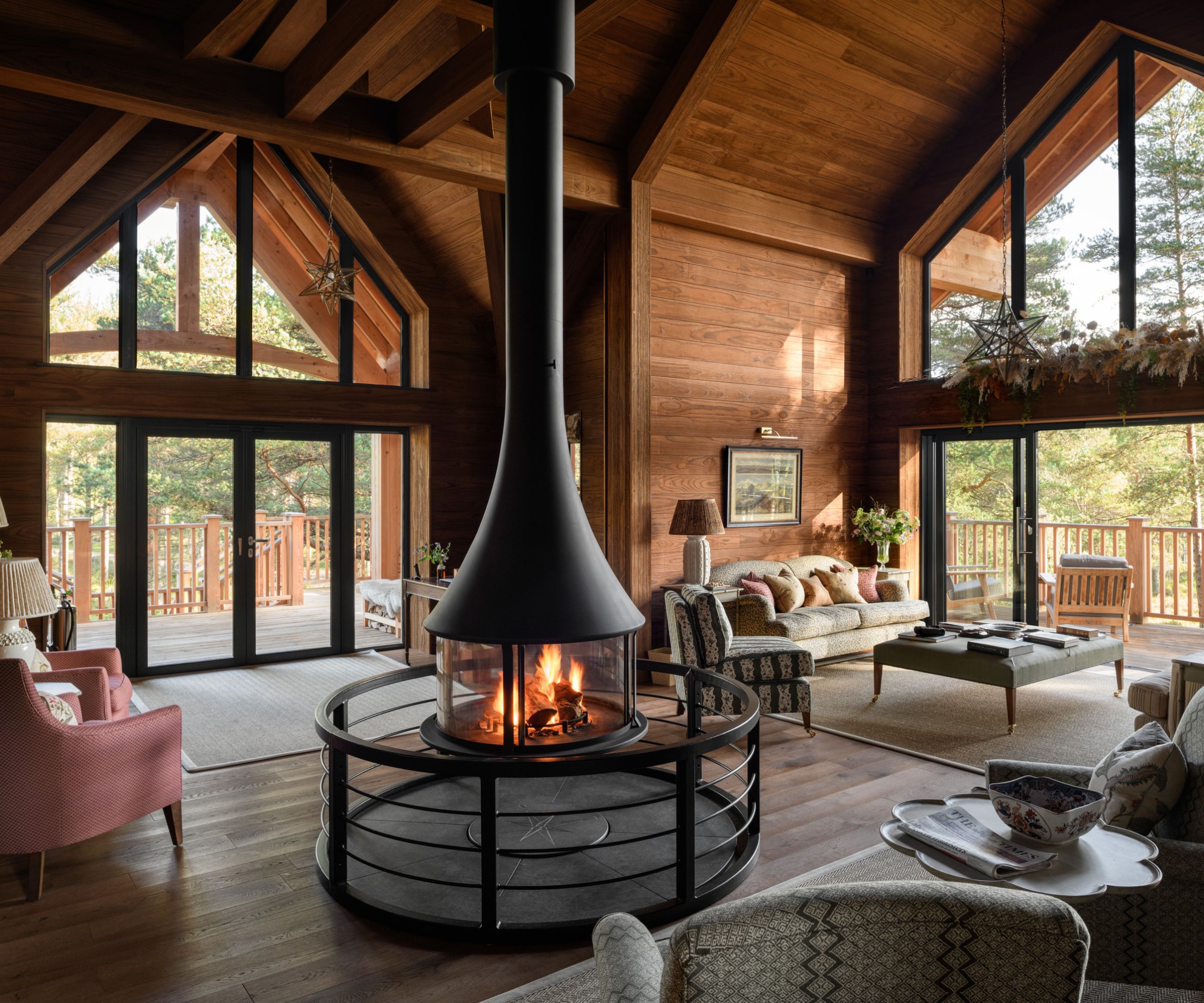 Interior of a timber-clad woodland lodge with soaring vaulted ceilings and full-height windows. A statement central fireplace anchors the open-plan living space, with layered textures, rustic wood finishes and forest views all around.