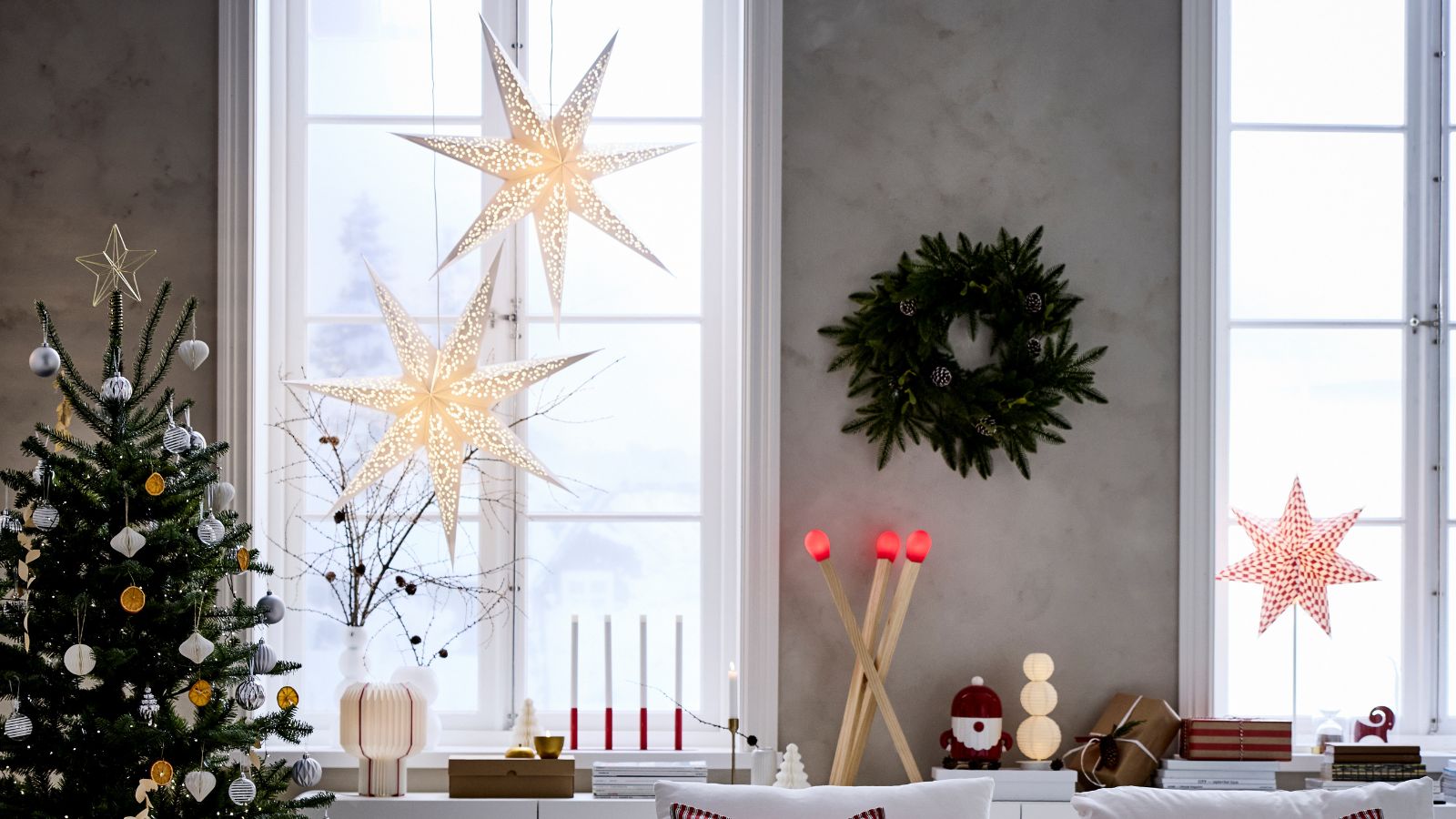 IKEA paper stars hung at the window of a living room with gray walls, a wreath between the window and a christmas tree decorated with white and gold