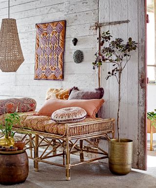 Boho lounge with cane bench, rattan pendant, large potted tree, textile wall hanging, and mixed patterned scatter pillows in warm, terracotta hues.