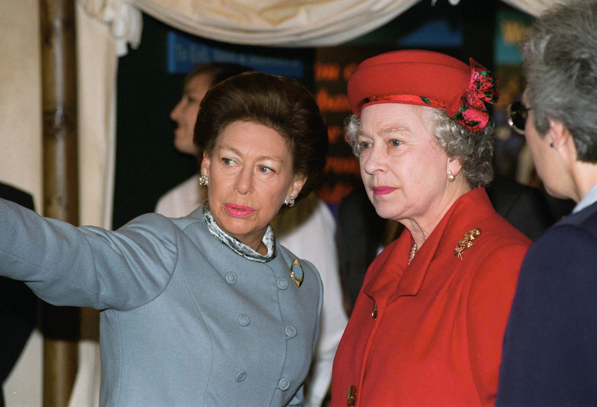 Princess Margaret in a blue coat pointing something out to Queen Elizabeth, dressed in a red hat and coat