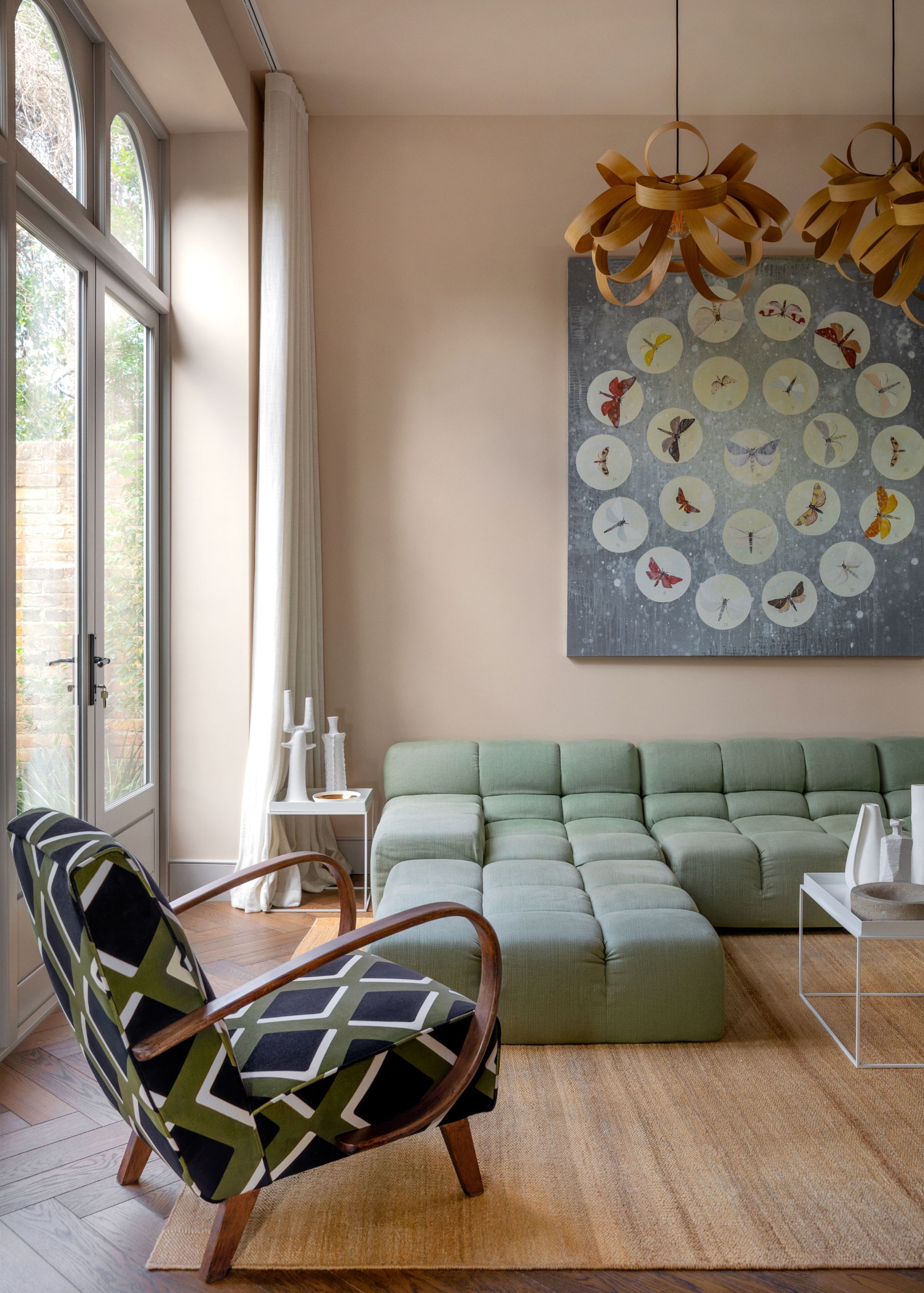 A modern living room with plaster pink walls, large glass doors, a light green minimal corner sofa, and a patterned accent chair.