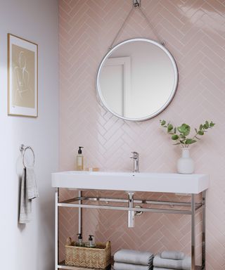 A small bathroom with light pink herringbone tiles, a silver circular mirror, a white freestanding sink, and a white wall with a beige wall art print and gray towel hanging from it