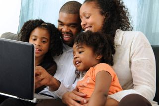 Mother and father hold two girls on their laps while viewing a laptop computer.