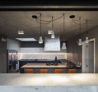 View of the kitchen. Countertops go all around the kitchen, continuing to the kitchen island with high metal chairs in orange. The walls are grey concrete, with white tiles and kitchen cabinets. The fridge is to the left.