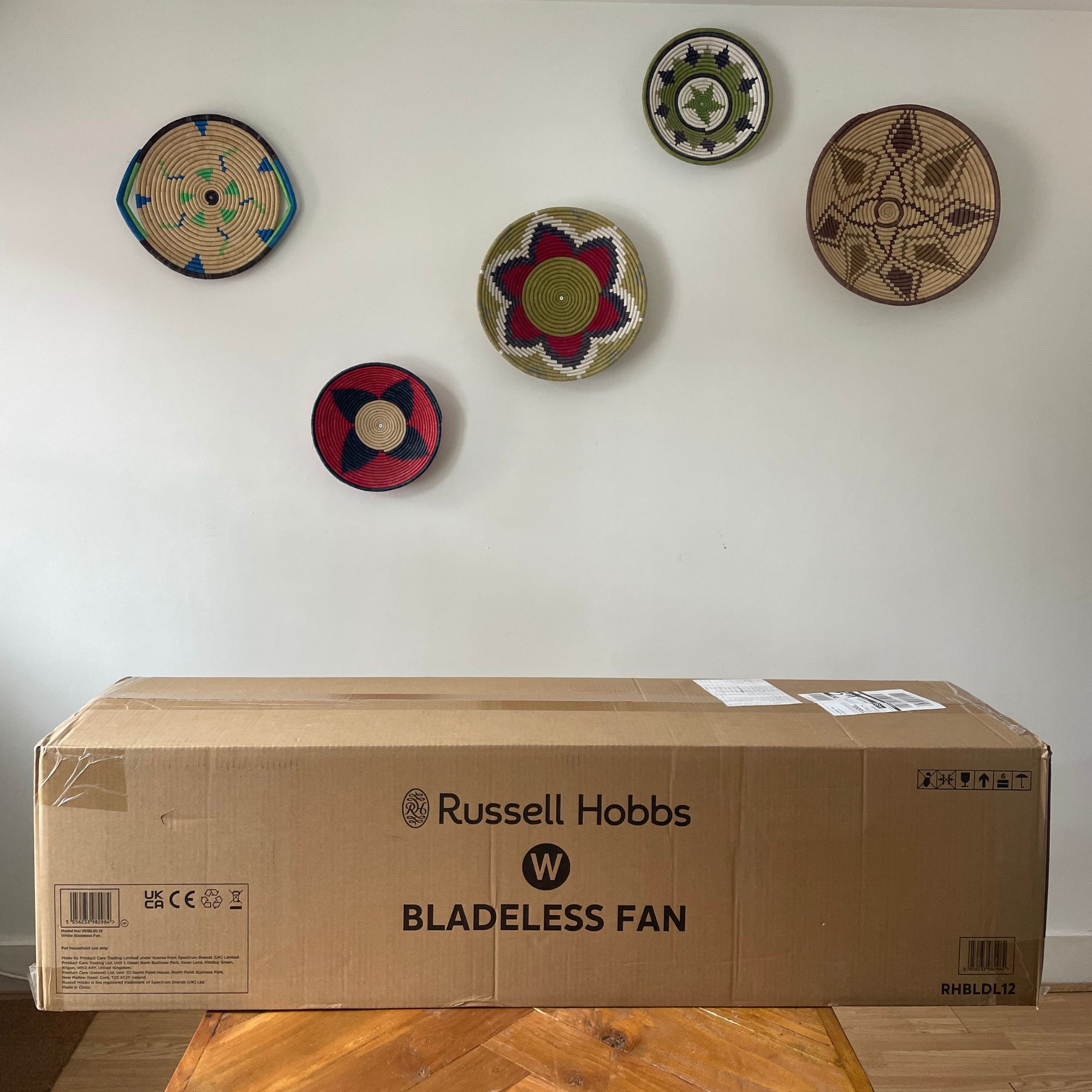 Russell Hobbs RHBLDL12 Bladeless Tower Fan box on a wooden table in front of a white wall with colourful baskets on it