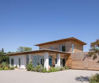 Large contemporary self build, clad in timber and render, with single storey entrance. The build is pictured against a blue sky on a sunny day.