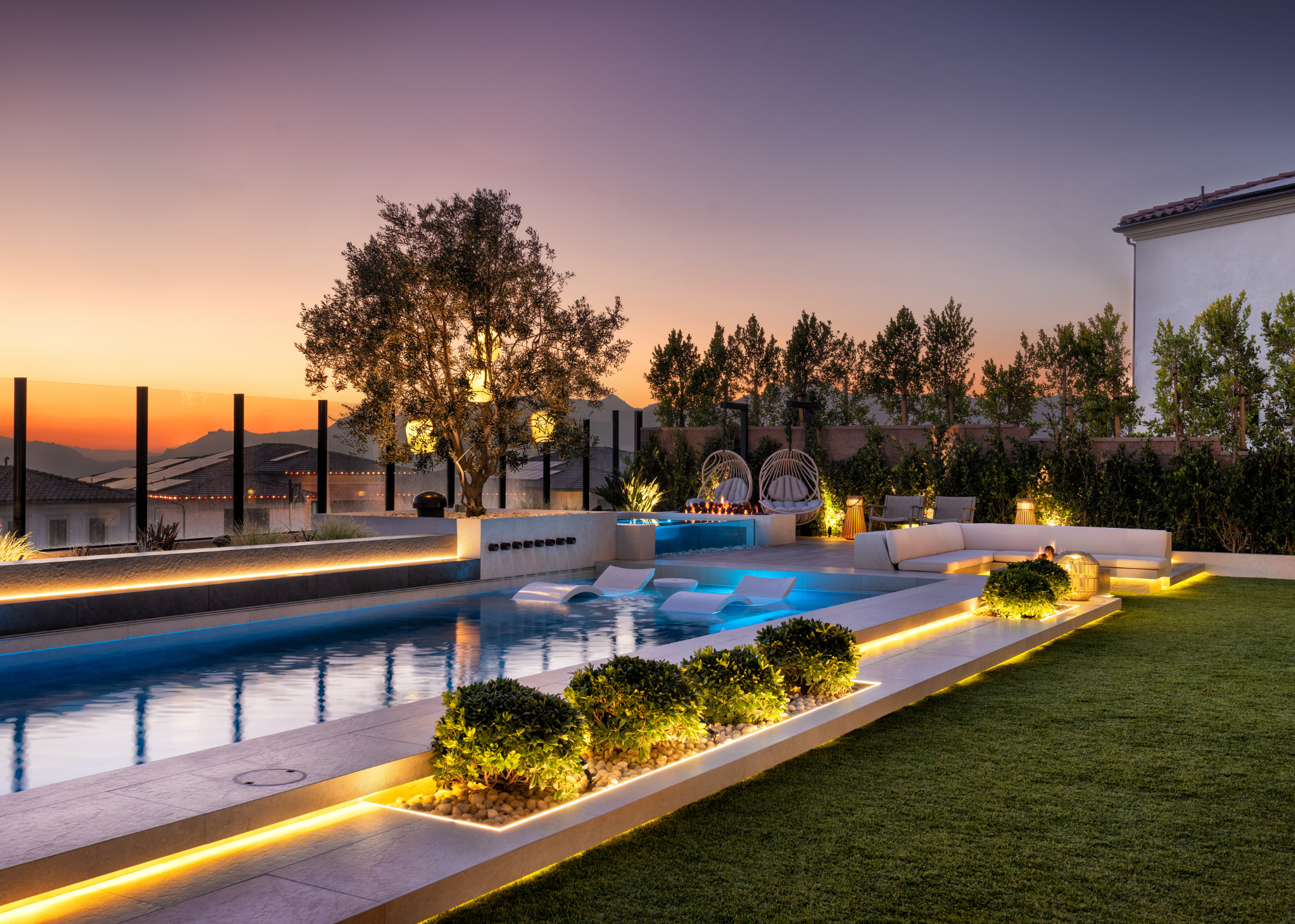 A modern california-style garden at dusk with a swimming pool and a warm, layered lighting scheme all around - from pendant lighting on the tree to lanterns and and strip lighting around the pool and flower beds