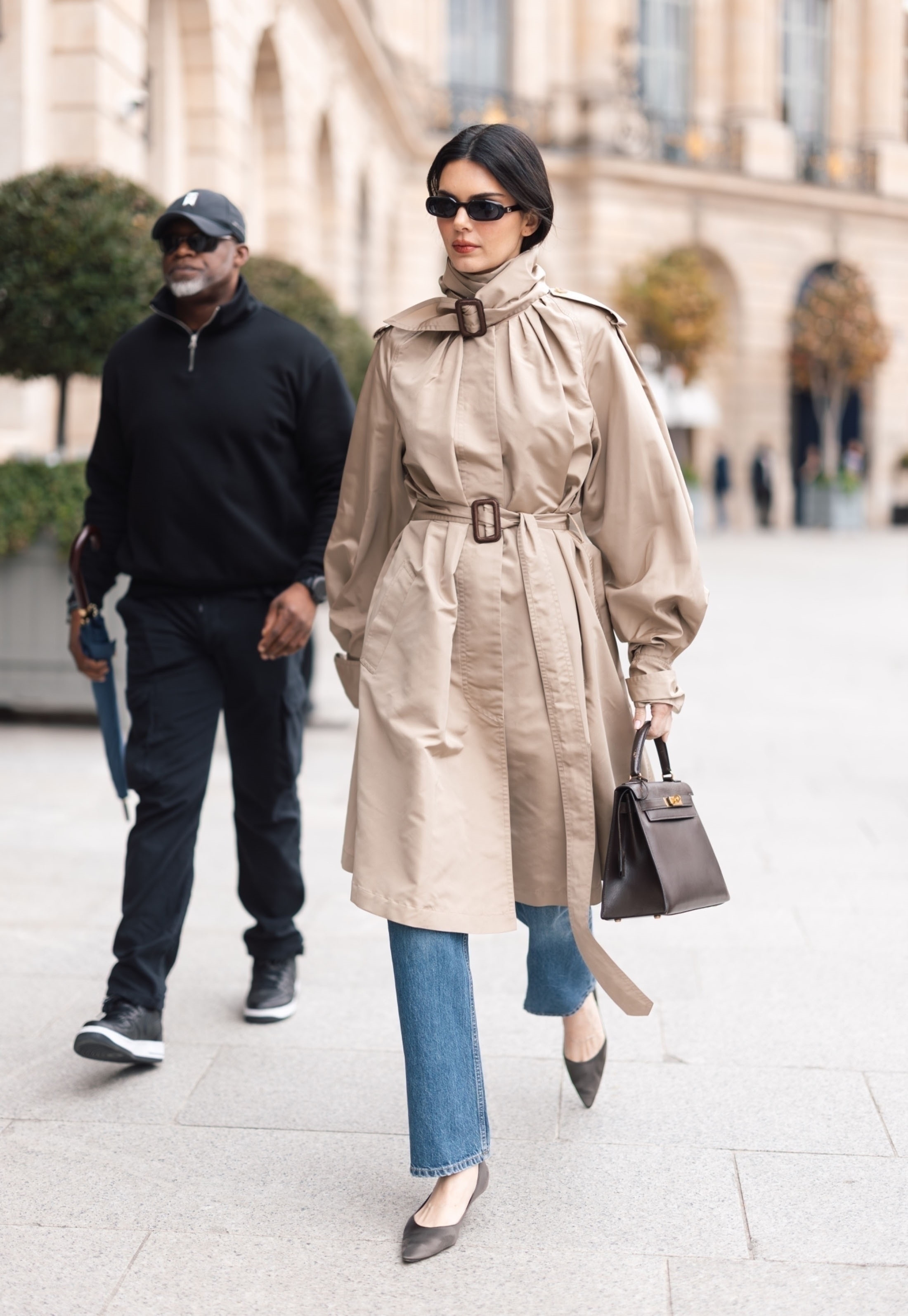 Kendall Jenner wearing a tan funnel-neck trench coat, blue jeans, and pointed toe black flats.