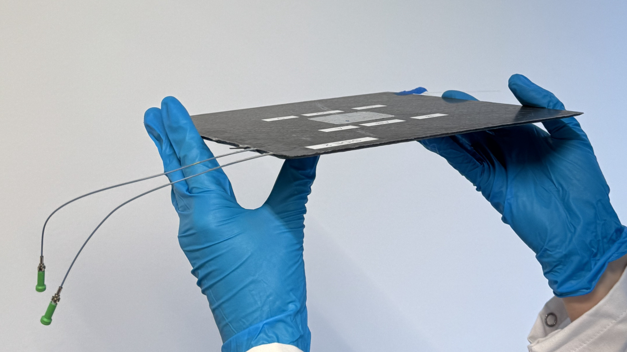 two blue-gloved hands hold up a wafer-thin black square about 16 inches on a side, which has two long wires trailing from it