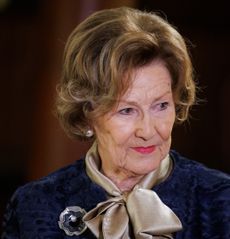 A headshot of Queen Sonja of Norway wearing a blue jacket and tie-neck blouse