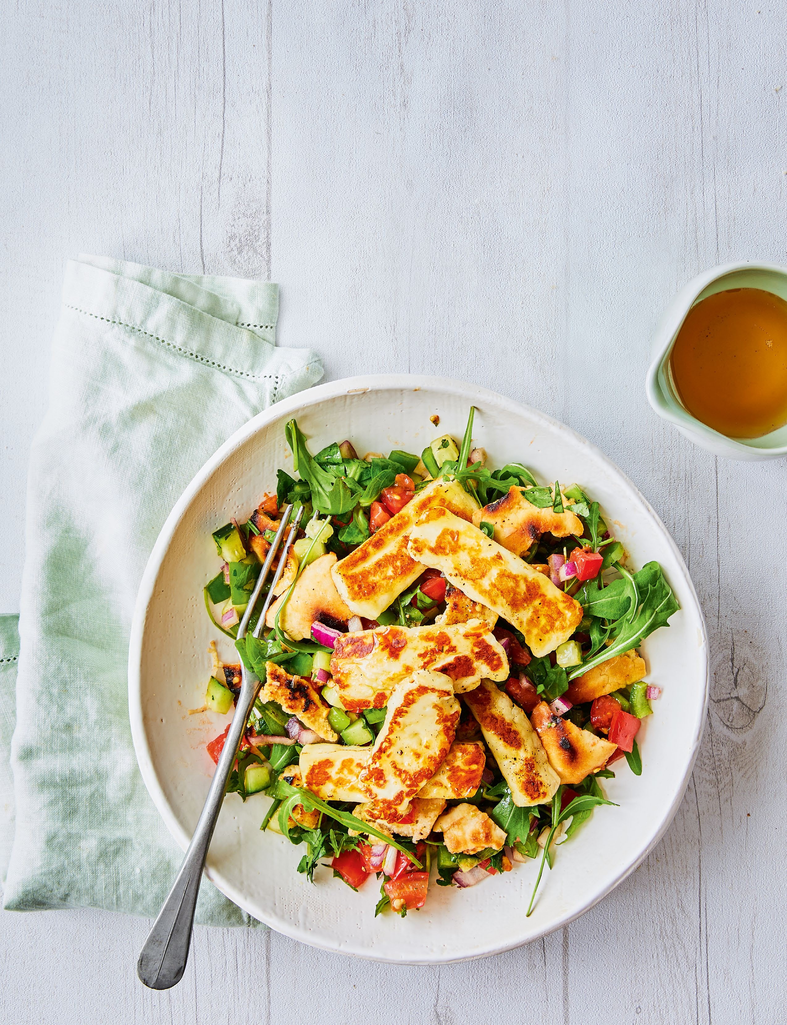 A vibrant Middle Eastern-inspired salad featuring grilled halloumi cheese, mixed greens, and colorful vegetables, served in a white bowl alongside a small cup of dressing.