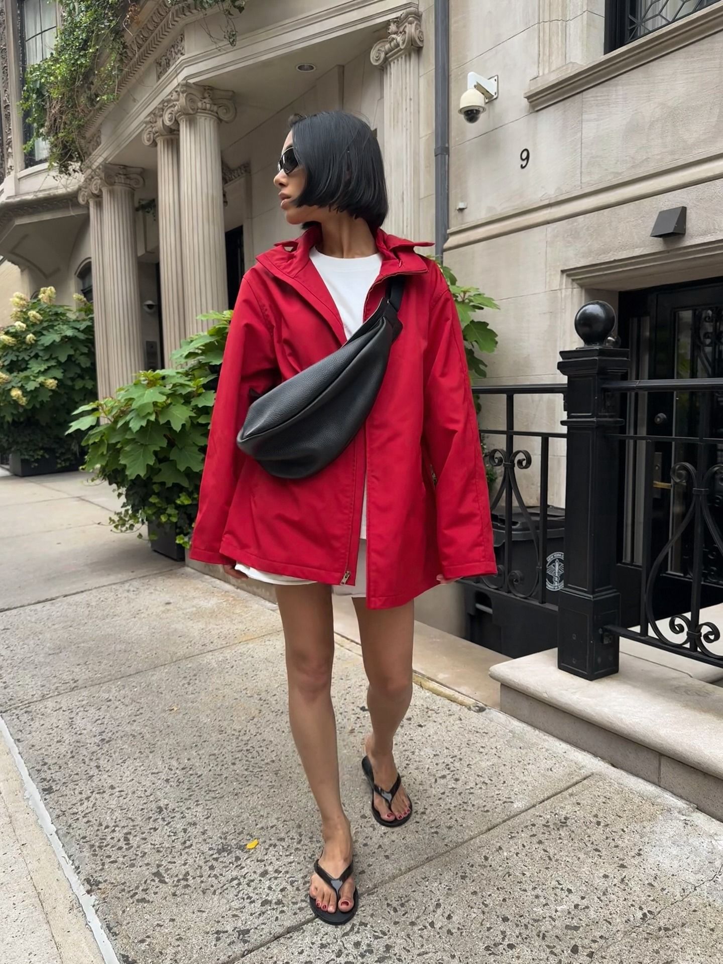 Woman wearing red windbreaker, black bag, white t-shirt, white shorts, and black flip flops.