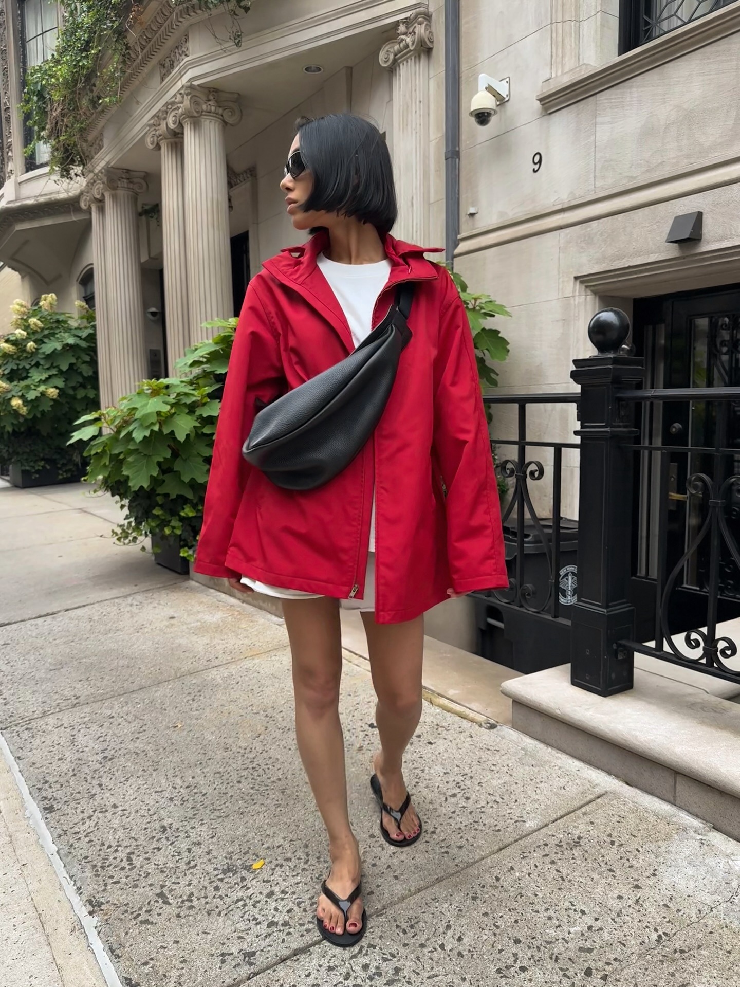 Woman wearing red windbreaker, black bag, white t-shirt, white shorts, and black flip flops.