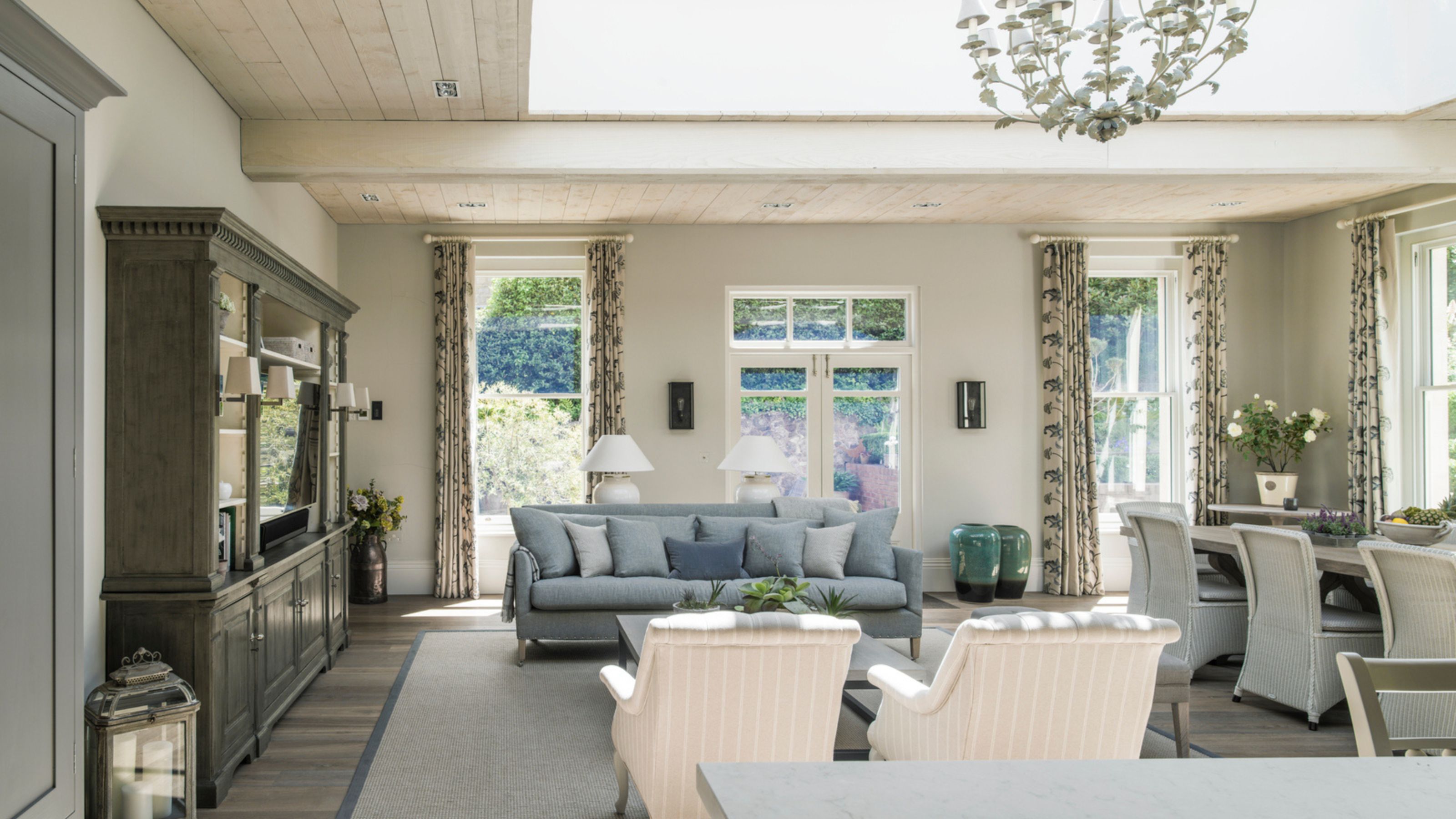 Bright dining/ living space with white walls, large windows with floral curtains, a white dining table and chairs, blue sofa, two pink and white striped chairs, and a large grey and glass cabinet.