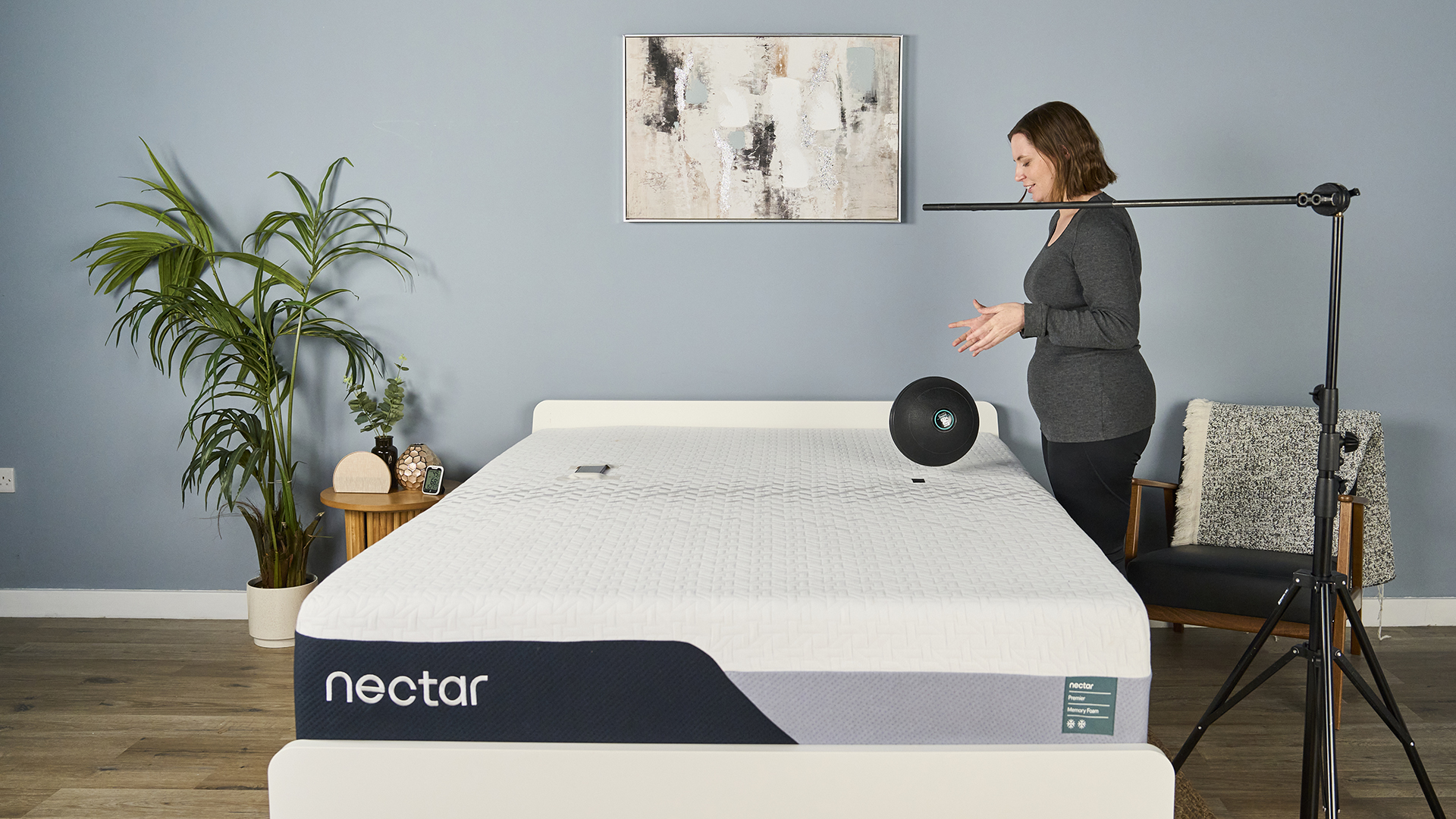 This image shows our sleep tester Ruth Jones during the slam ball drop test. She stands to the right of the Nectar Premier Memory Foam Mattress on a bed frame in our sleep studio and has just dropped a 5kg slam ball onto the marked drop zone on the mattress. To her right, a rig is set up to indicate 24 inches above the mattress; it's from this distance she dropped the ball.